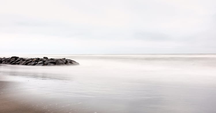 Langzeitbelichtung eines ruhigen Meeres. Ein steinerner Wellenbrecher ragt links ins neblige, weichgezeichnete Wasser.