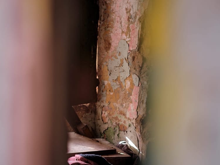 Blick durch einen unscharfen vertikalen Spalt in einen verfallenen Raum mit abblätternder Wandfarbe. Auf dem Boden liegt eine staubige Matratze mit Schutt und Stoff.