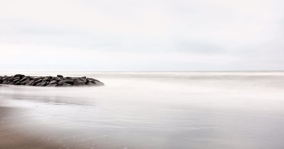 Langzeitbelichtung eines ruhigen Meeres. Ein steinerner Wellenbrecher ragt links ins neblige, weichgezeichnete Wasser.